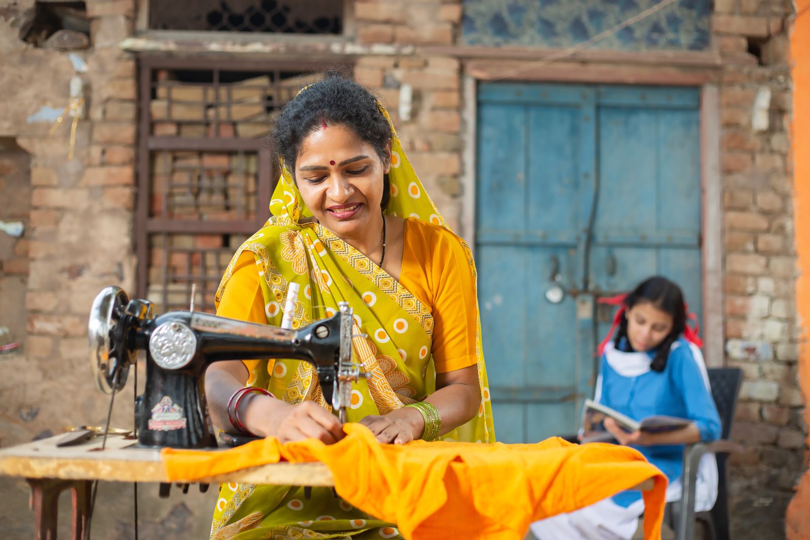 A woman sews at her machine outside her home while her daughter studies behind her