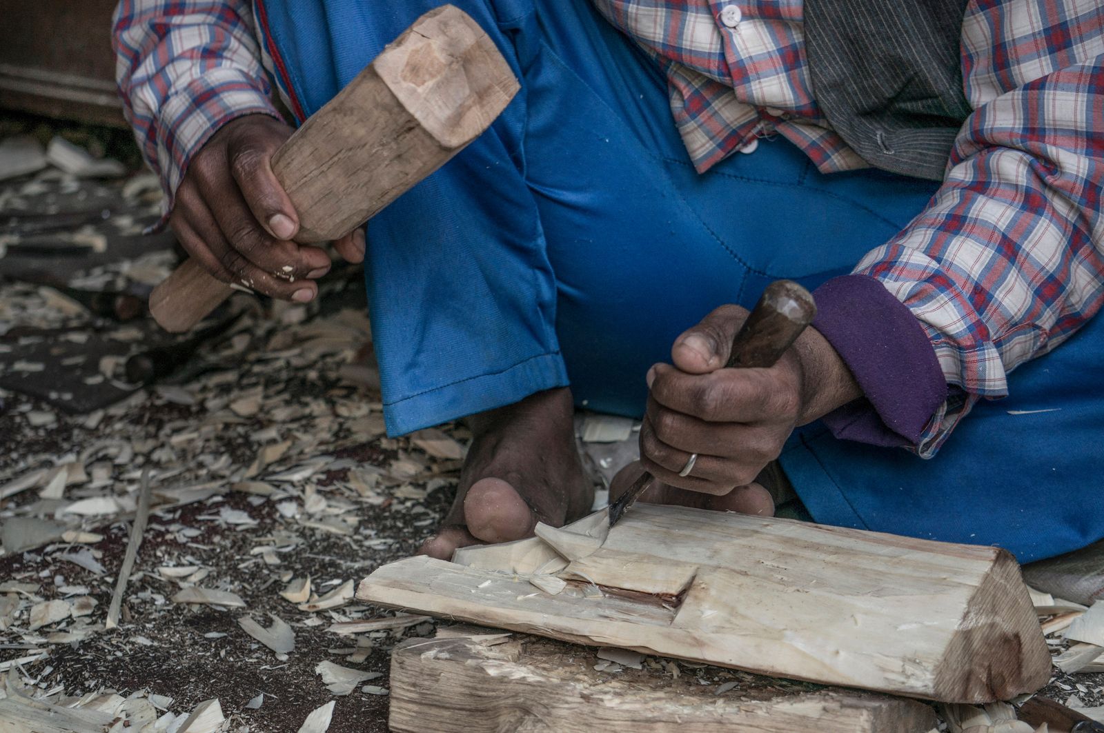 A craftsman's hands carve wood with a mallet and chisel, surrounded by wood shavings